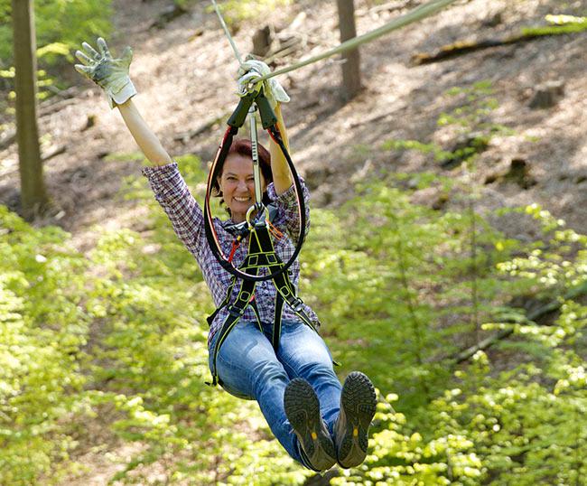 Lachende Frau rutscht mit ihrer angelegten Zipline-Ausrüstung am Seil entlang durch den Pfälzer Wald